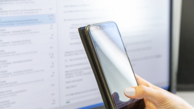 Hand hält Handy. Im Hintergrund ein Computermonitor